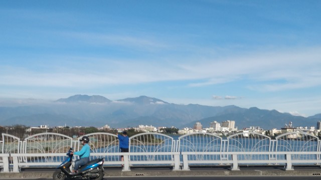 從東港進德大橋拍的照片，前面有一個人騎機車跟路人，背景有一些大樓跟山
不知道為什麼拍得有一點點彎彎曲曲