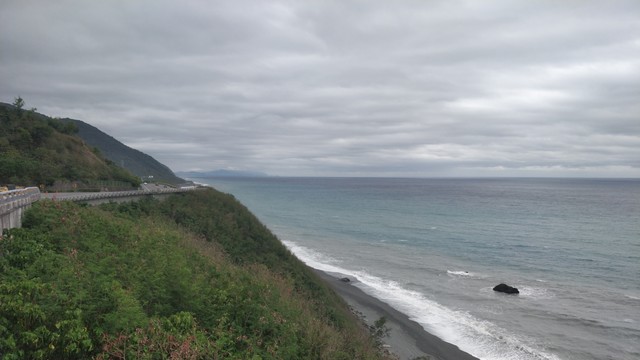 從南迴公路高一點的地方拍的照片，陰天，左邊有山跟公路，右邊有海跟小黑沙灘。海有三層，前面是灰色，中間是青綠色，後面是深藍色。最後面有很遠的山的側影。