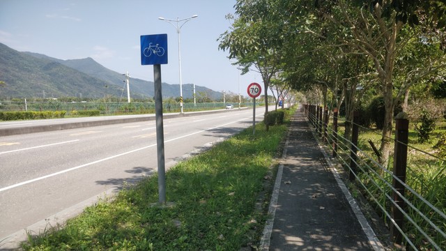 從旁邊的自行車道拍的台9線的照片。天氣很好，後面有山，在公路旁邊有自行車的藍牌子跟限速70的牌子，在自行車道旁邊有綠樹成蔭