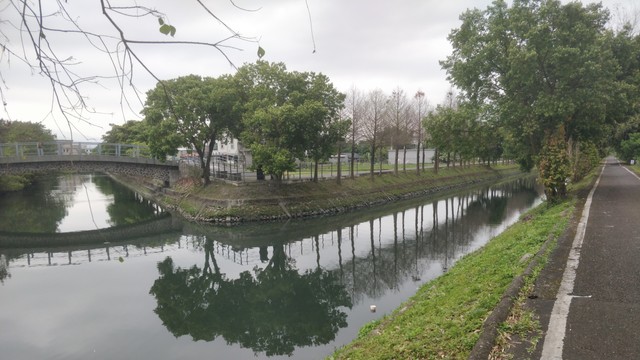 在羅東從沿運河的自行車道拍的照片。陰天，右邊有很直的自行車道，左邊有一座小橋，在中間河分開兩線，河旁邊有一些樹，樹跟橋倒影在河的水面上