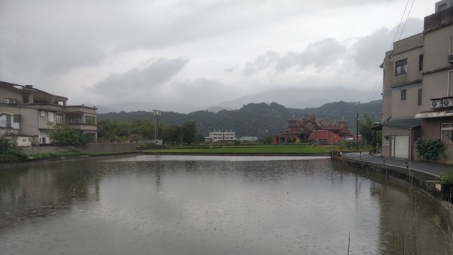 在礁溪鄉四結拍的照片，天氣下雨，前面有一個沒有植物的稻田，在它的兩邊有房子右邊還有一條路。後面有其他綠色的稻田跟一個紅色的寺廟，最後面有山丘。