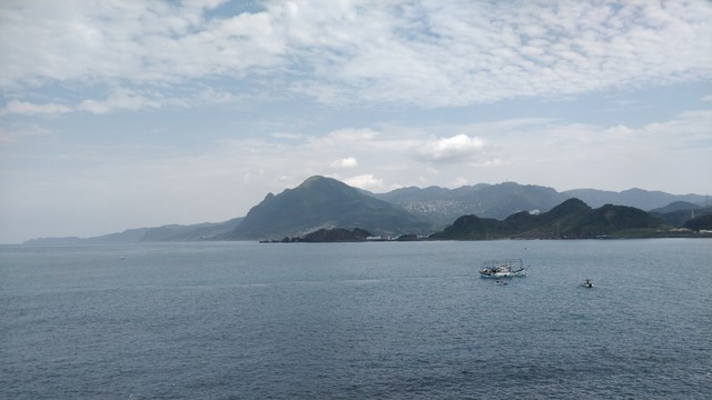 從八斗子拍的照片。天氣多雲，前面有藍色的海（番仔澳灣），海上有幾個船過去，後面有海岸跟山。山裡面有很遠的九份山城。